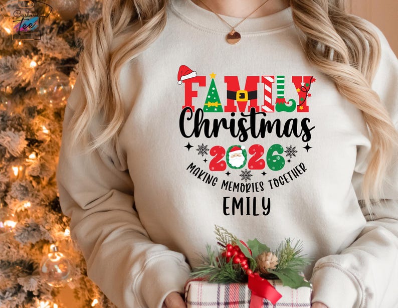 May include: A cream-coloured sweatshirt featuring a "Family Christmas 2026" design. The design includes a Santa hat, Christmas tree, and elf shoe. The text "Making Memories Together" and a name are also featured. A gift box with a red bow is in the foreground.