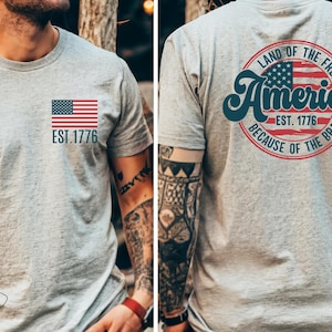 May include: A gray t-shirt with a small American flag on the front and a larger circle design on the back. The circle design features a larger American flag, the text "Land of the Free", "America", "Est. 1776", and "Because of the Brave".