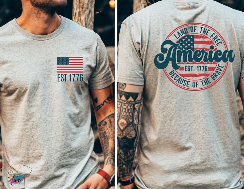 May include: A gray t-shirt with a small American flag on the front and a larger circle design on the back. The circle design features a larger American flag, the text "Land of the Free", "America", "Est. 1776", and "Because of the Brave".