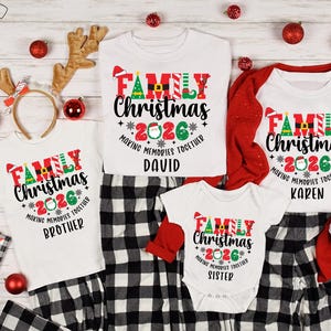 May include: A set of white Christmas tops and a babygrow, each with "FAMILY Christmas 2026" in red, green, and black. They are displayed with black and white checked pyjama bottoms. Christmas decorations are scattered around.