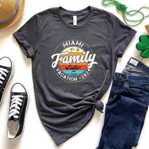May include: A dark grey t-shirt with a vintage-style circle graphic that says "Miami Family Vacation 2026" with the word "Sister" in a smaller font inside the circle. The graphic has a sunset color scheme with orange, yellow, blue, and green.