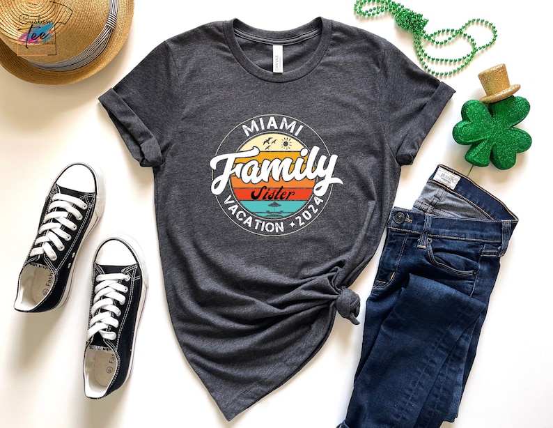 May include: A dark grey t-shirt with a vintage-style circle graphic that says "Miami Family Vacation 2026" with the word "Sister" in a smaller font inside the circle. The graphic has a sunset color scheme with orange, yellow, blue, and green.