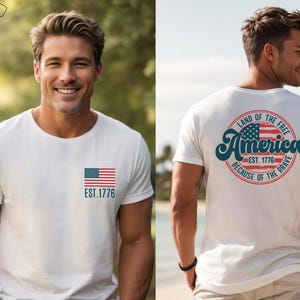 May include: A gray t-shirt with a small American flag on the front and a larger circle design on the back. The circle design features a larger American flag, the text "Land of the Free", "America", "Est. 1776", and "Because of the Brave".