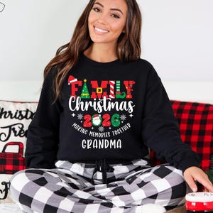 May include: Black sweatshirt with the text "FAMILY CHRISTMAS 2026" in red, green, and white. The sweatshirt is worn with black and white check pajama bottoms. A pillow with a red truck design is in the background.