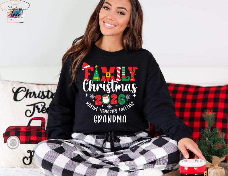 May include: Black sweatshirt with the text "FAMILY CHRISTMAS 2026" in red, green, and white. The sweatshirt is worn with black and white check pajama bottoms. A pillow with a red truck design is in the background.