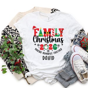 May include: A white t-shirt with the words "FAMILY Christmas 2026" in a festive design. The shirt includes a Santa hat, Christmas tree, and elf shoe graphics. The name "DAVID" is printed below the text. The shirt is surrounded by holiday decorations.
