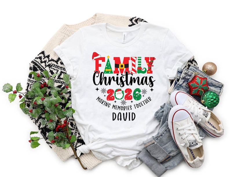 May include: A white t-shirt with the words "FAMILY Christmas 2026" in a festive design. The shirt includes a Santa hat, Christmas tree, and elf shoe graphics. The name "DAVID" is printed below the text. The shirt is surrounded by holiday decorations.