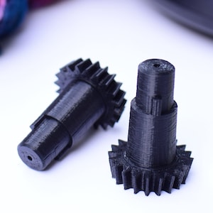 May include: Two black plastic gears with a star-shaped design. The gears are 3D printed and have a textured surface.