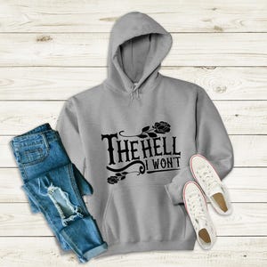 Op de afbeelding: Een grijs hoodie met de tekst "The Hell I Won't" in zwarte letters. De tekst is gestileerd met een bloemmotief en een zwarte roos.