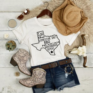 May include: White t-shirt with a black outline of the state of Texas. The text "I'm on the Lonestar Diet BBQ Mexican Whataburger Repeat" is inside the outline of the state. The shirt is laying on a white wooden surface with a straw hat, a pair of sunglasses, a small potted plant, a candle, and a pair of brown snake skin boots.