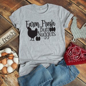 Op de afbeelding: Een grijs T-shirt met de tekst "Farm Fresh Butt Nuggets" en een zwart silhouet van een kip en eieren. Het shirt ligt op een houten oppervlak met een mand vol eieren, een rode bandana en een metalen haanfiguur.