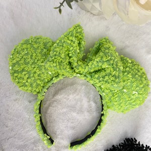May include: A lime green sequined headband with large bow-shaped ears. The headband is black and the ears are covered in small, shiny sequins. The accessory is designed to be worn on the head.