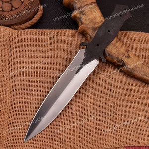 May include: A silver dagger with a black handle and a wooden sheath. The dagger is lying on a brown burlap surface.