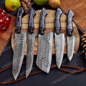May include: Set of five kitchen knives with black handles and Damascus steel blades. The blades have a wavy pattern and are arranged on a wooden cutting board.
