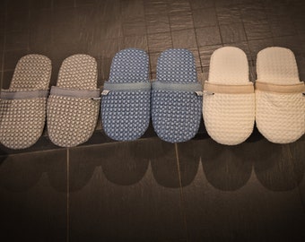 Könnte beinhalten: Sechs Paar Hausschuhe in drei verschiedenen Farben: Grau, Blau und Weiß. Die Hausschuhe sind aus einem weichen, strukturierten Stoff gefertigt und haben ein schlichtes, bequemes Design.