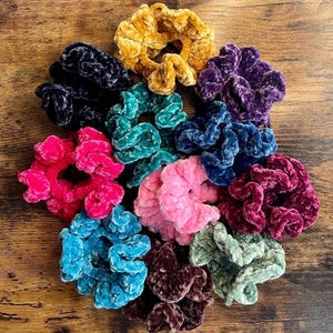 May include: A collection of colorful velvet scrunchies in various shades, including pink, blue, purple, and gold. These hair accessories are arranged on a wooden surface, showcasing their soft texture and vibrant hues.