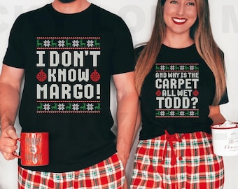 May include: Matching black t-shirts with holiday-themed text. One shirt reads "I DON'T KNOW MARGO!" and the other says "AND WHY IS THE CARPET ALL WET TODD?" Both shirts have a festive pattern. The couple is also wearing red plaid pajama pants.