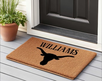 May include: A rectangular brown doormat with the name "WILLIAMS" in black, above a black longhorn silhouette. The mat is placed on a gray wooden porch, near a black door and a potted plant.