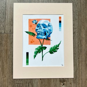 May include: A blue skull with a white flower growing from its top, set against a peach background. The skull is surrounded by green leaves. The image includes a color palette with shades of green, blue, and peach.