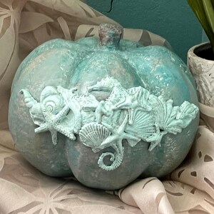 May include: A teal-colored pumpkin with a textured surface, adorned with a decorative band of seashells and starfish. The pumpkin is placed on a patterned fabric, with a potted plant in the background.