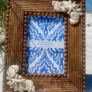 May include: A wooden picture frame with a white and blue patterned design inside. The frame is decorated with white seashells on the corners. The frame is 4 x 6 inches or 10 x 15 cm. The text on the frame reads "Handcrafted in India, Fabriqué à la main en Inde, 4 x 6 / 10 x 15cm".