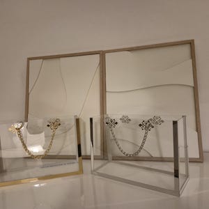 Könnte beinhalten: Zwei transparente Acryl-Handtaschen mit Kettenhenkeln. Eine hat goldfarbene Metallverzierungen, die andere silberfarbene. Dahinter befinden sich zwei gerahmte abstrakte Kunstwerke in neutralen Farben.