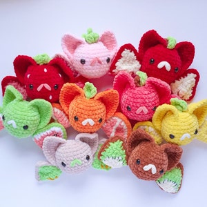 May include: A collection of colorful, crocheted fruit-themed plush toys. The toys are shaped like bats, with fruit slices as wings and various fruits as the heads, including strawberries, oranges, and watermelons. The toys are in shades of red, pink, green, and yellow.