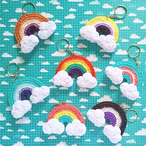 May include: A collection of handmade crochet rainbow keychains with fluffy white cloud accents. Each keychain features a different color scheme, including orange, yellow, red, teal, and purple. The keychains are attached to silver-colored key rings.