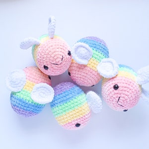 May include: Five handmade crochet bee toys in pastel rainbow colors. Each bee has white wings, black eyes, and a smiling mouth. The toys are made of soft yarn and are arranged on a white surface.