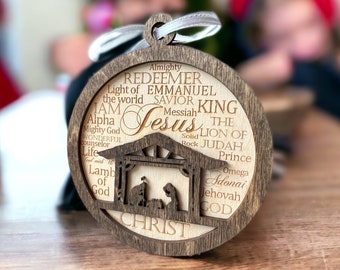 May include: A wooden Christmas ornament with a nativity scene cutout and the words "Almighty Redeemer Emmanuel Light of the world Savior King I AM Messiah Jesus The Lion of Judah Prince Omega Adonai Jehovah God Christ" engraved around the edge.