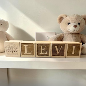 May include: Wooden blocks spelling "LEVI" with a light wood finish and dark wood inlay. One block features an engraved image of Noah's Ark. Two teddy bears and a small card are in the background.