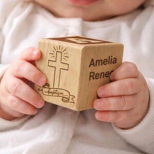 May include: A wooden block engraved with a cross and the word "baptism" on a banner. The block also features the name "Amelia Rener". The block is held by a baby.