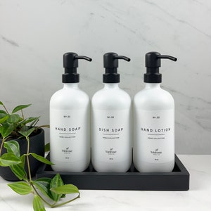Op de afbeelding: Drie witte pompflacons met zwarte pompen en etiketten. De flacons zijn gelabeld met "No. 01 Handzeep", "No. 02 Afwasmiddel" en "No. 03 Handlotion". De flacons staan op een zwarte dienblad.