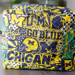 May include: A quilted, rectangular cosmetic bag featuring a repeating pattern of yellow and navy blue text and graphics. The text includes phrases like "GO BLUE" and "MICHIGAN". The bag has a zipper closure and a diamond-stitched pattern.