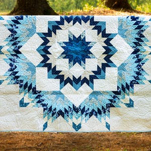 May include: A handmade quilt with a starburst design in shades of blue and white. The quilt features intricate geometric patterns and is displayed outdoors, showcasing its craftsmanship and color palette. The quilt is approximately 60 inches by 70 inches.