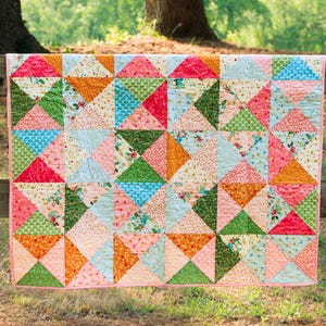 May include: A colorful patchwork quilt with a geometric design. The quilt features triangles in shades of pink, orange, green, blue, and white. The quilt is hanging on a wooden fence, with a pink border.