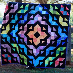 May include: A colorful quilt with a geometric design. The quilt features a black background with a central starburst pattern in shades of orange, pink, blue, green, and purple. The quilt is bordered with a blue binding.