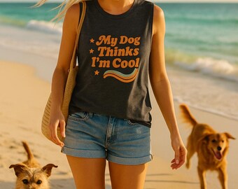 May include: A woman wearing a dark gray tank top with the text "My Dog Thinks I'm Cool" in orange and blue. She is walking on a sandy beach with two dogs. She is wearing denim shorts and sunglasses.