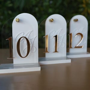 May include: Three white table number signs with gold numbers 10, 11, and 12. Each sign has a clear acrylic backing and a gold-colored stud. The numbers are in a bold, elegant font, perfect for weddings or events.