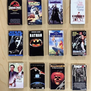 May include: A collection of twelve VHS tapes featuring movie titles like "Jurassic Park," "Back to the Future," "Star Wars," and "Batman." The tapes are arranged on a light wood surface, showcasing classic film artwork.