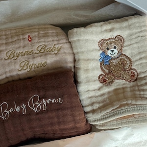 May include: A set of baby items, including two beige and one brown muslin cloths, and a folded beige towel. The cloths are embroidered with the words "Byrne Baby Byrne" and "Baby Byrne". The towel features a teddy bear with a blue bow.