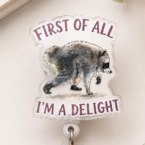May include: A clear acrylic badge reel featuring a watercolor illustration of a raccoon. The text "FIRST OF ALL" is printed in dark purple above the raccoon, and "I'M A DELIGHT" is printed in dark purple below the raccoon. The badge reel has a retractable cord and a metal clip.
