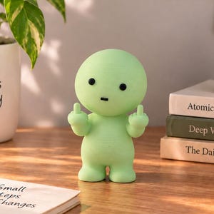 May include: A light green, rounded figurine with a smooth surface, two small black eyes, and a small black mouth. The figurine is standing upright with both arms raised, each hand extending a middle finger in a gesture of defiance.