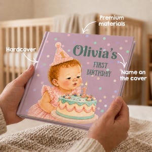 May include: A hardcover book with a light purple cover featuring a pattern of colorful dots. The cover displays an illustration of a child in a party hat blowing out a candle on a cake, with the text Olivia's FIRST BIRTHDAY printed in dark green.