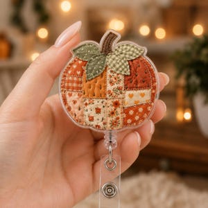 May include: A pumpkin-shaped badge reel featuring a patchwork design with various fabric patterns in shades of orange, red, and cream. The pumpkin has green gingham and solid green fabric leaves at the top and a brown stem, all encased in clear plastic.