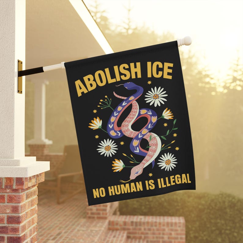 May include: Black flag with the words "ABOLISH ICE" and "NO HUMAN IS ILLEGAL" in yellow. A colorful snake design is in the center, with white daisies and yellow flowers. The flag is hanging outside on a white pole.