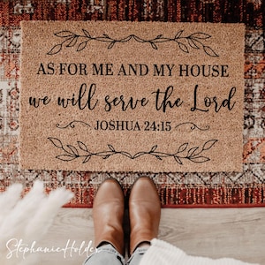 Könnte beinhalten: Eine rechteckige Fußmatte mit dem Text "AS FOR ME AND MY HOUSE we will serve the Lord JOSHUA 24:15" in schwarzer Schrift. Die Matte hat eine natürliche beige Farbe mit einem dekorativen Blätterrand. Braune Stiefel sind unten zu sehen.