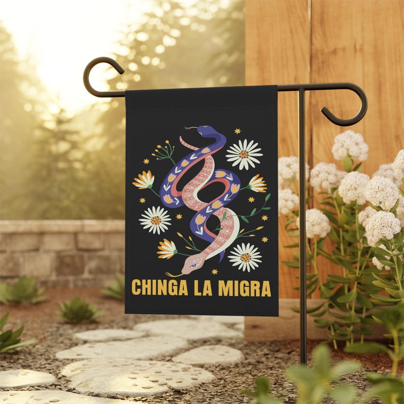 May include: Black garden flag featuring a colorful illustration of a snake with floral accents and the text "CHINGA LA MIGRA" in gold. The snake is pink, blue, and white, surrounded by white and yellow flowers. The flag is displayed outdoors.