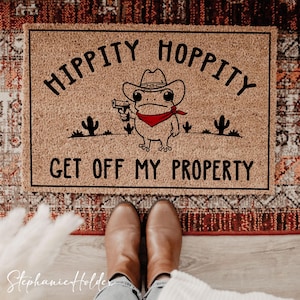 Może przedstawiać: Prostokątna wycieraczka z napisem "HIPPITY HOPPITY GET OFF MY PROPERTY" i kreskówkową żabą w kapeluszu kowbojskim i trzymającą pistolet. Mata jest brązowa z czarną obwódką i wzorem. Dwa małe kaktusy po obu stronach żaby.