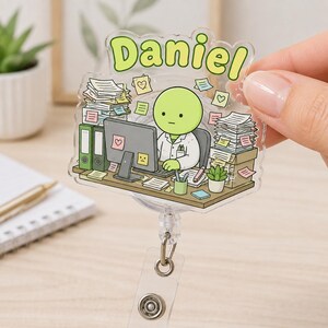 May include: A clear acrylic badge reel featuring a cartoon illustration of a green character sitting at a desk with stacks of paper, a computer, and a potted plant. The name Daniel is printed in large light green letters at the top of the badge.